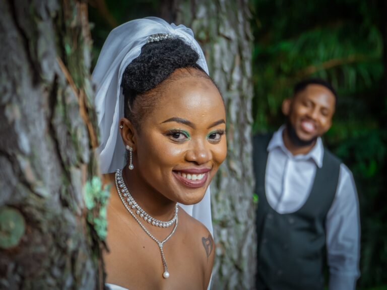 Bride smiling beside a tree with her groom behind her