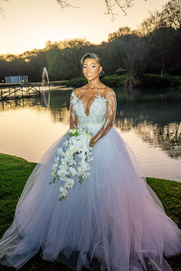 Bride in a full gown holding an orchid bouquet beside a lake at sunset