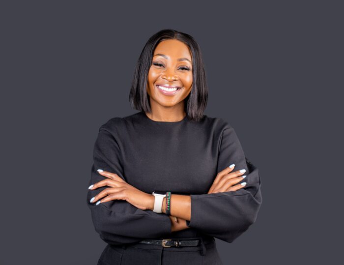 Professional headshot, black top, arms crossed, bright smile on a charcoal backdrop, by Studio24.7
