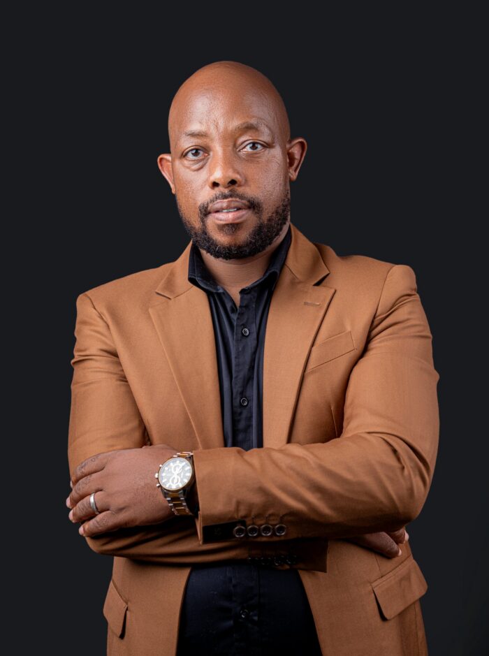 Professional headshot, tan blazer, arms crossed, on a dark backdrop, by Studio24.7